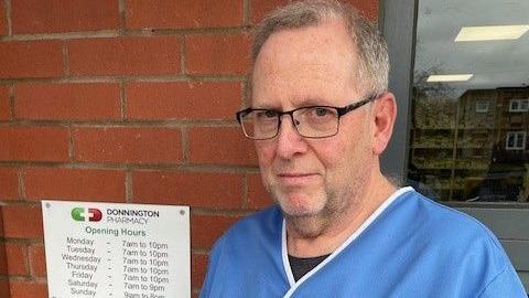 A man with black framed glasses and grey hair with a blue medical top on stood beside a brick wall with Donnington Pharmacy and opening hours written on a sign