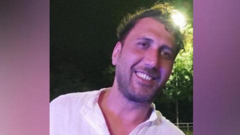 Mustafa Qays Obadey smiles while wearing a white shirt. He is posing at night with a street light seen shining behind his head.