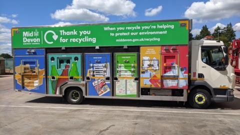 A Mid Devon District Council recycling lorry parked in a parking area. Blue and cloudy skies.