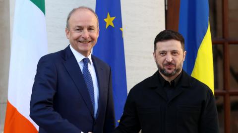 Taoiseach Micheál Martin and Ukrainian President Volodymyr Zelensky standing in front of their country's flags. Martin is wearing a blue suit and Zelensky is all in black.