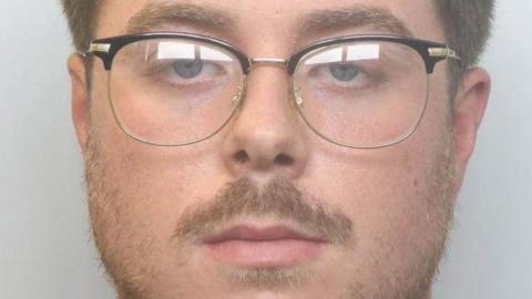 A man with short brown hair, a moustache and some facial hair and glasses is looking at the camera in a police mugshot.