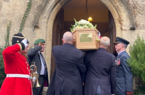 Men carry a coffin into a church flanked by men in military uniform who are saluting