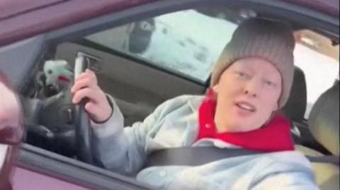 Image shows a woman wearing a blue and white jacket, a red hooded jumper and a beige hat. She is sitting in a red vehicle with the window open and her arm resting on the window frame