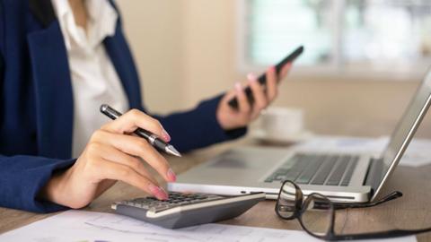 Close-up of a woman in a suit looking at her mobile and tapping on a calculator, with a laptop visible in the background