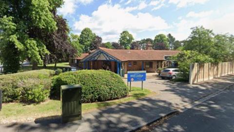 A single storey building with a car park, bordered by small hedges. A sign saying 'Mill Lodge' is at the entrance of the car park.