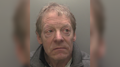 A police custody shot of David Glover. He has short grey hair and is wearing a grey collared jumper. He is looking to the right of the camera. The wall behind him is grey.