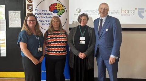 Three women and one man standing in front of a power point presentation. Behind them is a pull-up banner which reads Surrey Partnership for Inclusion of Neurodiversity in Schools.  