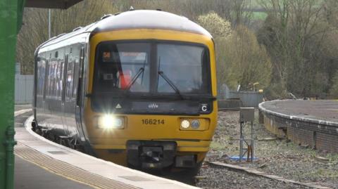 A GWR train pulling into a station and its platform with its front lights on. Its got a yellow front and green on the side. 