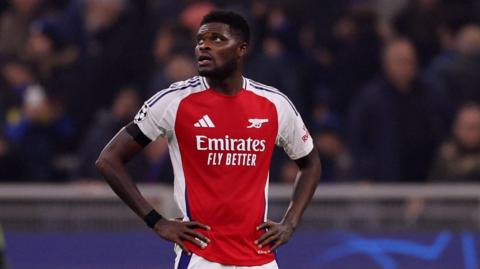 Thomas Partey in his football kit. He stands with his hands on his hips. A blurred crowd is behind him.