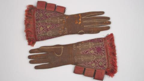 A pair of large brown leather gloves with red silk embroidery and gold embellishment, belonging to King James I, circa 1610s. They are on a grey background.