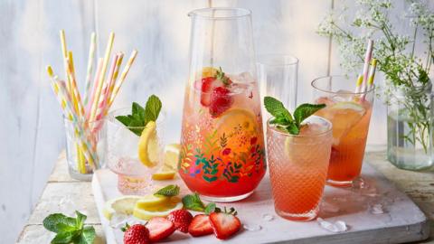 A printed jug full of strawberry lemonade on ice with strawberries and mint sits on a board with two glasses of lemonade and scattered slices of lemons and strawberries.