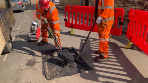 Two people in orange high-vis clothing putting tarmac mix in a square hole in a road. Bright orange barriers are to the right, with a small section of a lrry visible on the left
