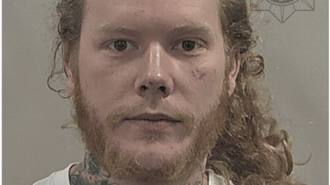 Police mugshot of a man with long, curly, unkempt, sandy-coloured hair tied back in a pony tail and a straggly ginger beard and moustache. He has a heavily tattooed neck and is wearing a white T-shirt.