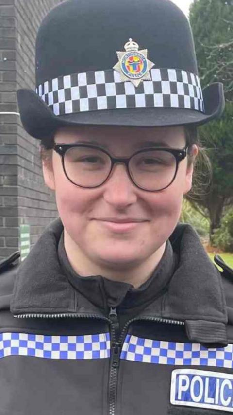 Jess Turnbull, wearing police uniform and smiling. She is standing in front of a dark grey brick building with a wooden entrance to one side. Behind her to the other side is a patch of grass, a tall, thin bush-like tree and some other trees, like you might find in the gardens of an office building. Jess is wearing a rounded police hat and black rimmed glasses and has her dark hair tied back.