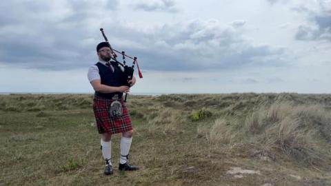 Ryan Fitzpatrick stands on the marram grass of the North Denes Beach in Great Yarmouth. He is wearing a black hat, white shirt, red tie, dark blue waistcoat, kilt and sporran, and white knee-length socks, with black shoes. He is holding, and playing his bagpipes.