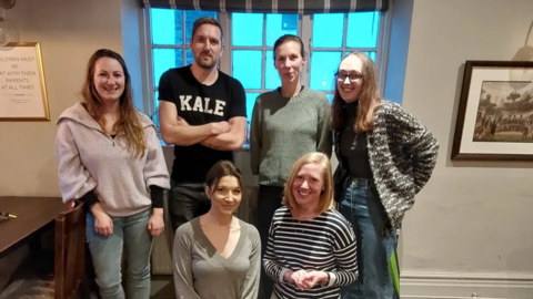 A group of six people pose for a photograph in a pub. The group consists of five females and one male.