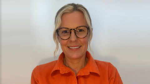 Swimming teacher Glynis Sheppard is smiling towards the camera. She has blonde hair which is tied up with some strands coming down and is wearing glasses. She is wearing an orange blouse. 