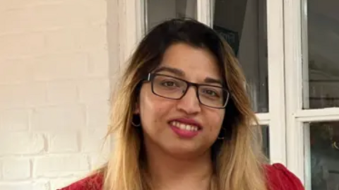 A close-up image of Lowestoft Mayor Nasima Begum. She is wearing glasses and a red t-shirt and is standing in front of a window. She is smiling into the camera.