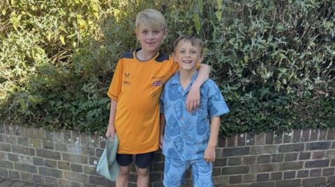 Two young boys standing side-by-side. A blonde boy in an orange shirt has his arm around a shorter boy with brown hair who is wearing a blue shirt.