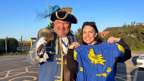 Bob Smitherman, wearing the tricorn hat and ceremonial robes of the Worthing Town Crier, stands with his bell on his shoulder, while Alison Ferns stands beside him, holding up a dress decorated with the Sussex coat of arms.