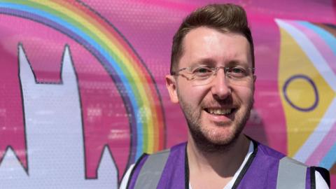 Arran Hart smiles as he stands in front of a pink Lincoln Pride banner, which features a rainbow flying over a white outline of Lincoln Cathedral. He is wearing a purple hi-vis vest and glasses.