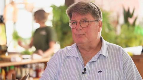Rob Stander, wearing a blue and white striped shirt and glasses. The background is blurred behind him. There is a woman sitting at a table in the background.