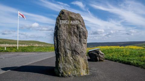 A large stone with England carved into it in a layby next to a road.