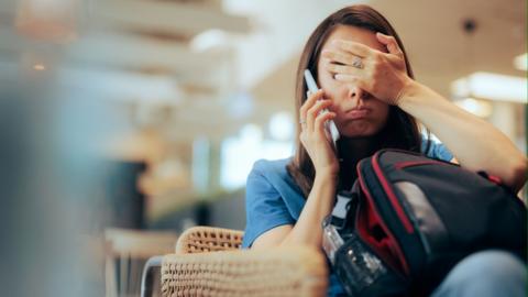 A woman with her hand over her face, talking on the phone while holding a bag in an airport lounge