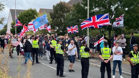 A group of people carrying Union Jacks and England flags in a suburban street. A line of police look on.