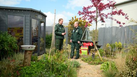Two ambulance crew members, both men, standing inside a garden with green shrubs and a pink-leaved tree and a grey outbuilding with glass windows.
