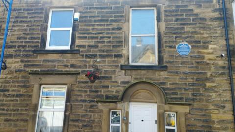 A picture of a terraces stone built house with a blue plaque high on the wall next to the upstairs window 