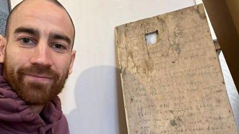 Tom Yuen smiles at the camera in front of the old panel, he has a brown -ish ginger beard and brown eyes. 