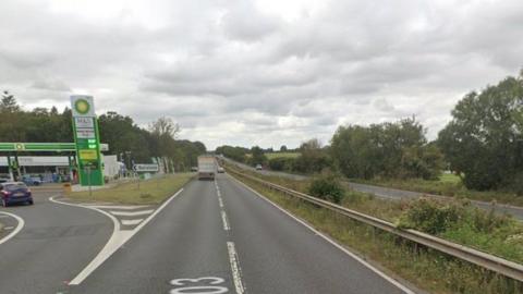 Roadside services on left - fuel station and coffee shop single carriageway road in centre, tree lined bank on right.