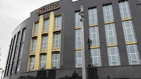 A large corner building restored to its original 1930s look. Black granite cladding cover the building which has a number of long windows. The centre of the building is painted gold.
