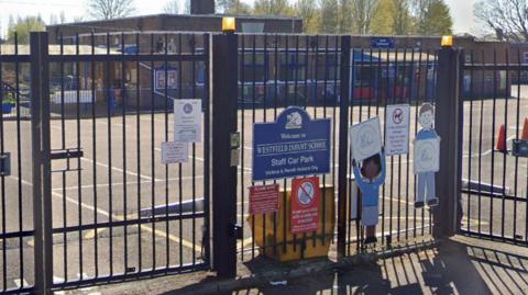 Outside of Westfield Infant School - a gate around the school is at the front of the photograph with signs on, and the playground and building is in the background. 