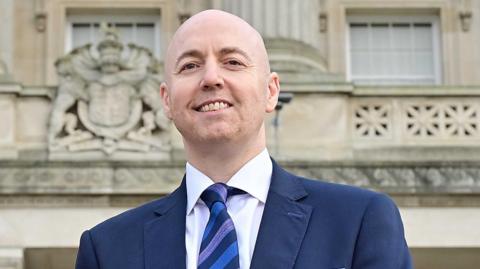 A man, Jon Burrows, is smiling at the camera. He is bald and is wearing a navy jacket over a white shirt and a blue and navy diagonally striped tie. He is standing outside of Parliament Buildings at Stormont in Belfast. 