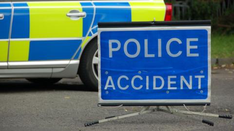 A sign reading POLICE ACCIDENT, with white letters on a blue background, that has been placed on a road. A police car is behind it.