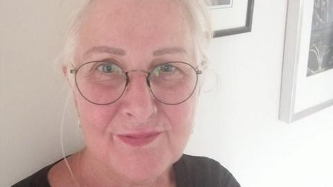A woman with white hair, tied back, wearing round black-rimmed spectacles and with a nose ring, smiles up at the camera as she takes a selfie. She is wearing a dark grey/black top and is standing against a white wall with framed pictures hanging on it.