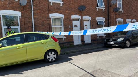 'Police Do Not Cross' Tape creates a barrier across a street of terraced houses, with a police officer behind a green car parked on the street. A black car is also parked on the street.