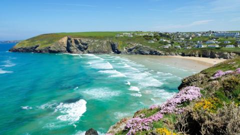 A wide shot of the bay at Mawgan Porth taken on a sunny day. The sea is a mixture of blues and greens with some white flecks on the waves. There is a small sliver of light sand beach visible between the cliffs of the bay and on a hillside running up above the beach are dozens of properties