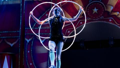 A circus performer holds up three hoops while smiling during a stage performance.