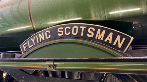 A green-painted metal cylinder with a curved black sign which has the words flying Scotsman written on it in gold lettering and some black-painted metalwork below