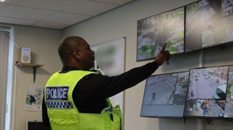 A police officer in uniform pointing to a bank of CCTV screens on a wall