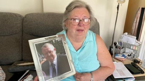 Catherine Curtis sat on a sofa, holding a framed photograph of Michael Curtis. She appears sad and as if she has been crying.