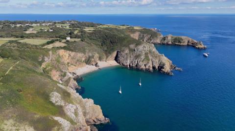 An aerial image of the island of Sark, showing a beach and cliff faces under sunny skies. There re a number of boats bobbing in the sea.
