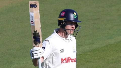 Glamorgan batter Ben Kellaway, wearing a helmet and batting gloves, raises his bat after reaching a milestone
