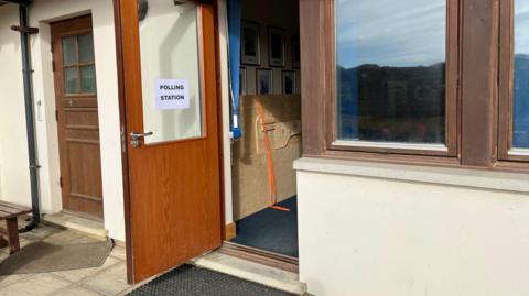 Sark's pollong station has an open wooden door with a glass panel on which there is a printed sign that reads POLLING STATION. There is another wood and glass door to the left and a wood-framed window to the right. Inside a folded wooden table can be seen and there are picture frames on the wall.