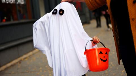 A small child wearing a ghost costume for Halloween. The white sheet costume completely covers them, with only two holes for the child to see. The child is trick or treating and is holding a pumpkin bucket for sweets.