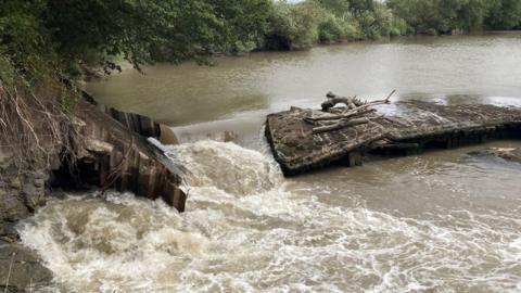 A photo showing a concrete weir across the River Severn. There is a large hole to the left side of the weir with water pouring through