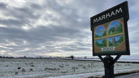 Marham village sign, with paintings of a plane, house, field and church. It stands next to a road and snowy fields, with sheep grazing.